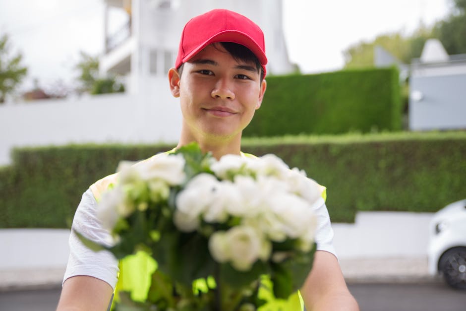 A young male flower delivery courier wearing a red cap and high-visibility vest is smiling as he presents a bouquet of fresh white roses and green foliage. The bouquet is held out in front of him, sli